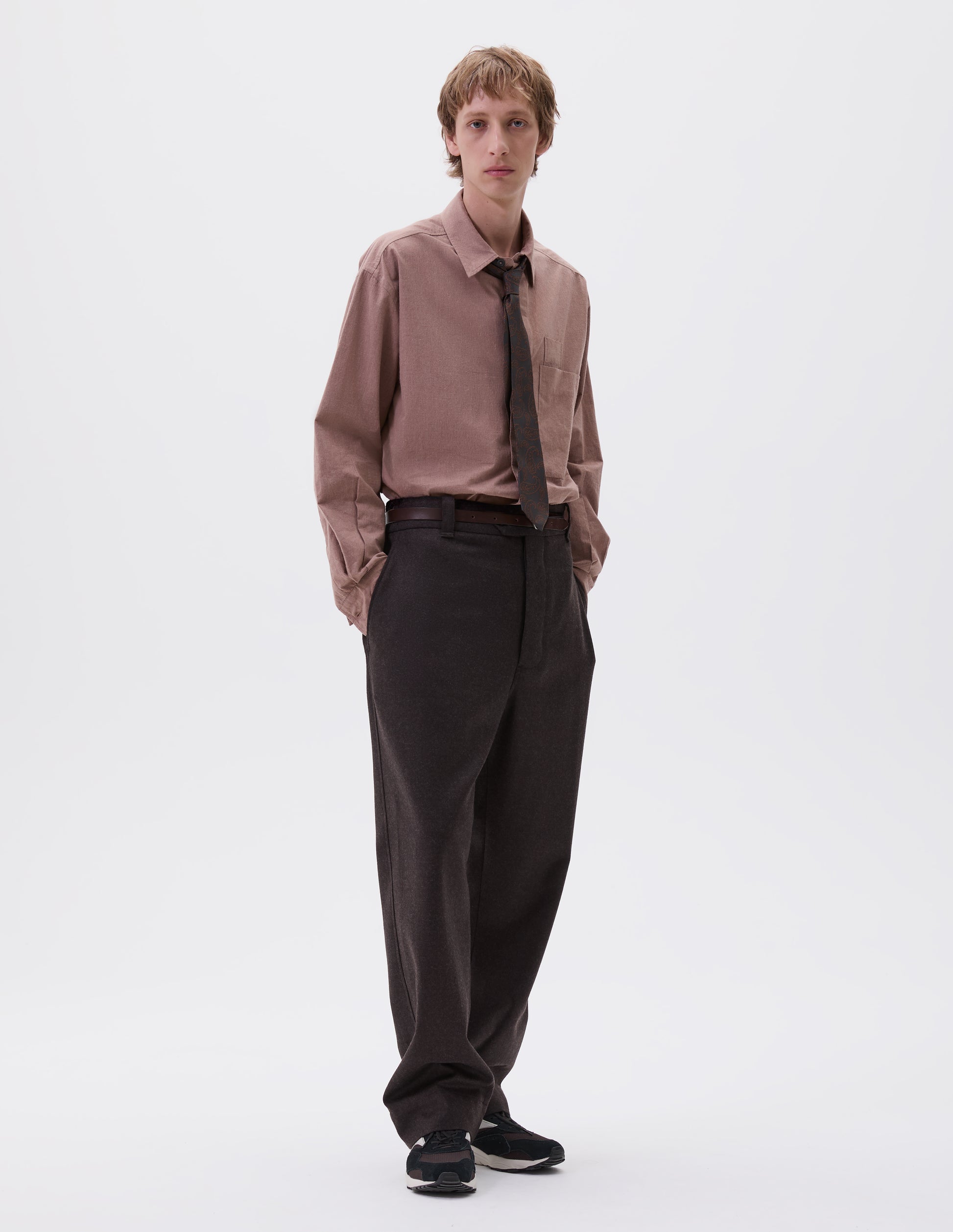 Full body image, front view of model wearing brown wool trousers, with half elasticated waistband, belt loops, horn button tab fastening and slant pockets. Styled with pink shirt, brown paisley tie, brown leather belt  and black and brown trainers.