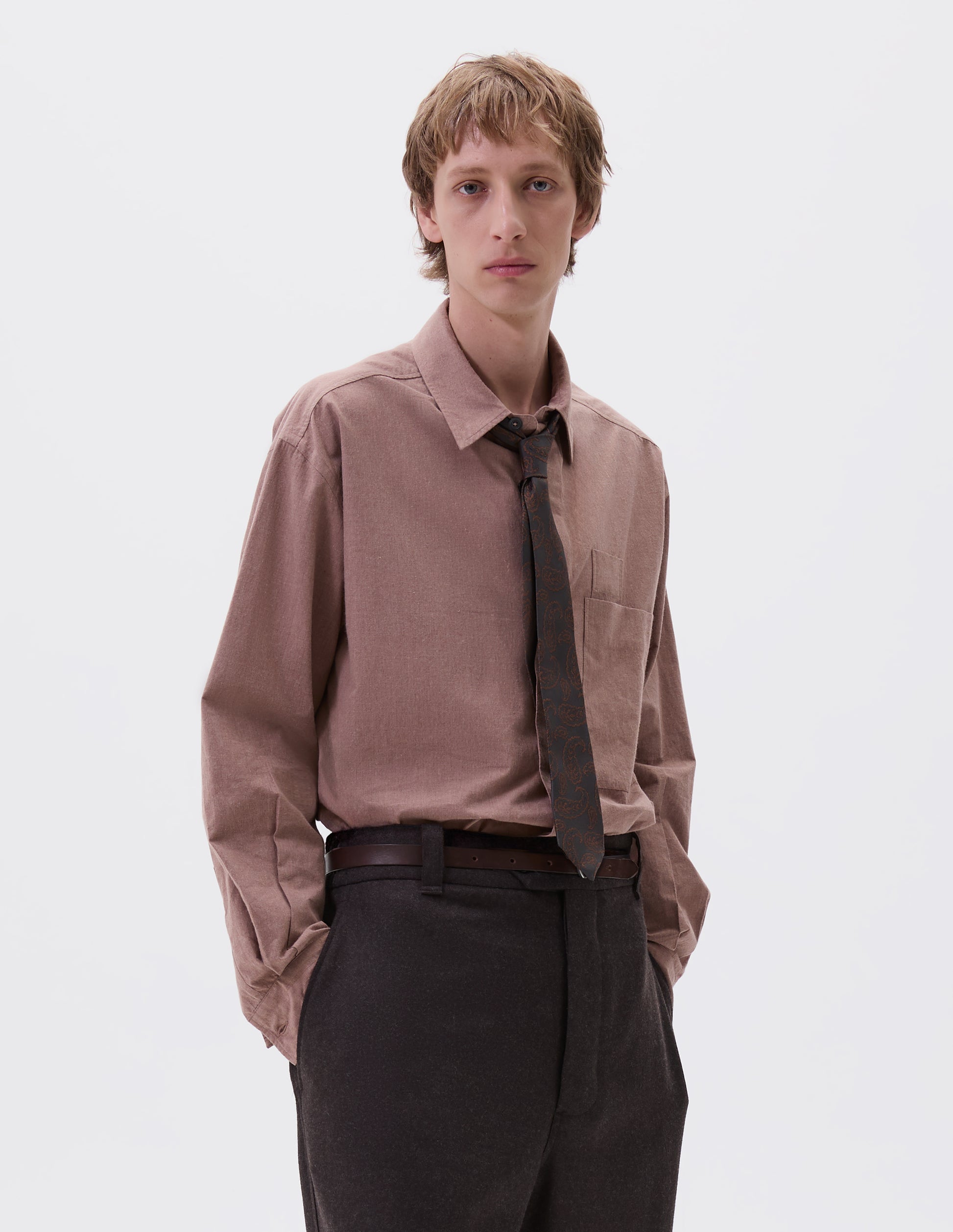Cropped body image, front view of male model wearing dark brown slim leather belt with sterling silver buckle and contrast hand tack detail. Styled with pink shirt, brown paisley tie and brown wool trousers. 