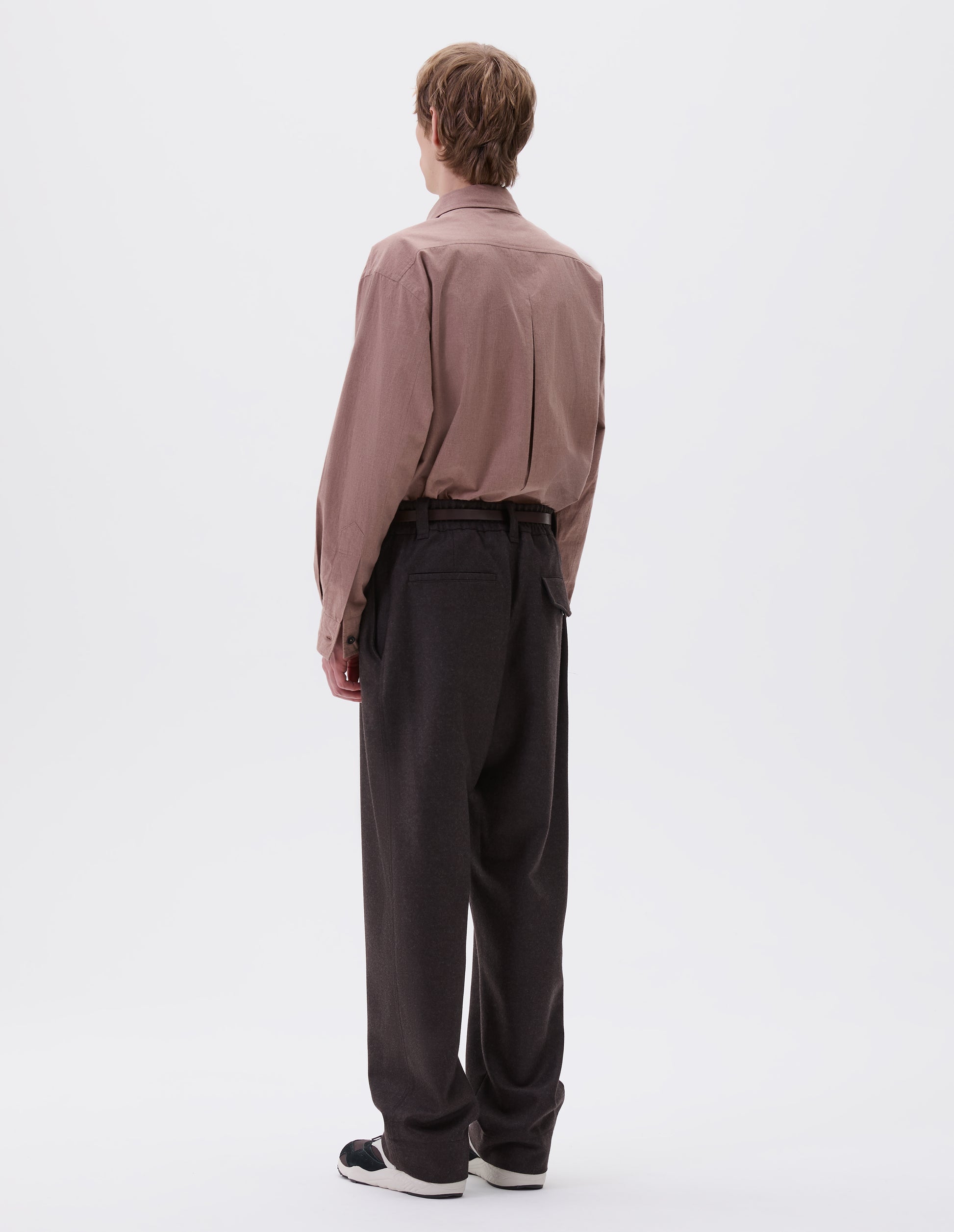Full body image, back view of model wearing brown wool trousers, with half elasticated waistband, belt loops and welt and concealed button fastening flap back pockets. Styled with pink shirt, brown paisley tie, brown leather belt  and black and brown trainers.