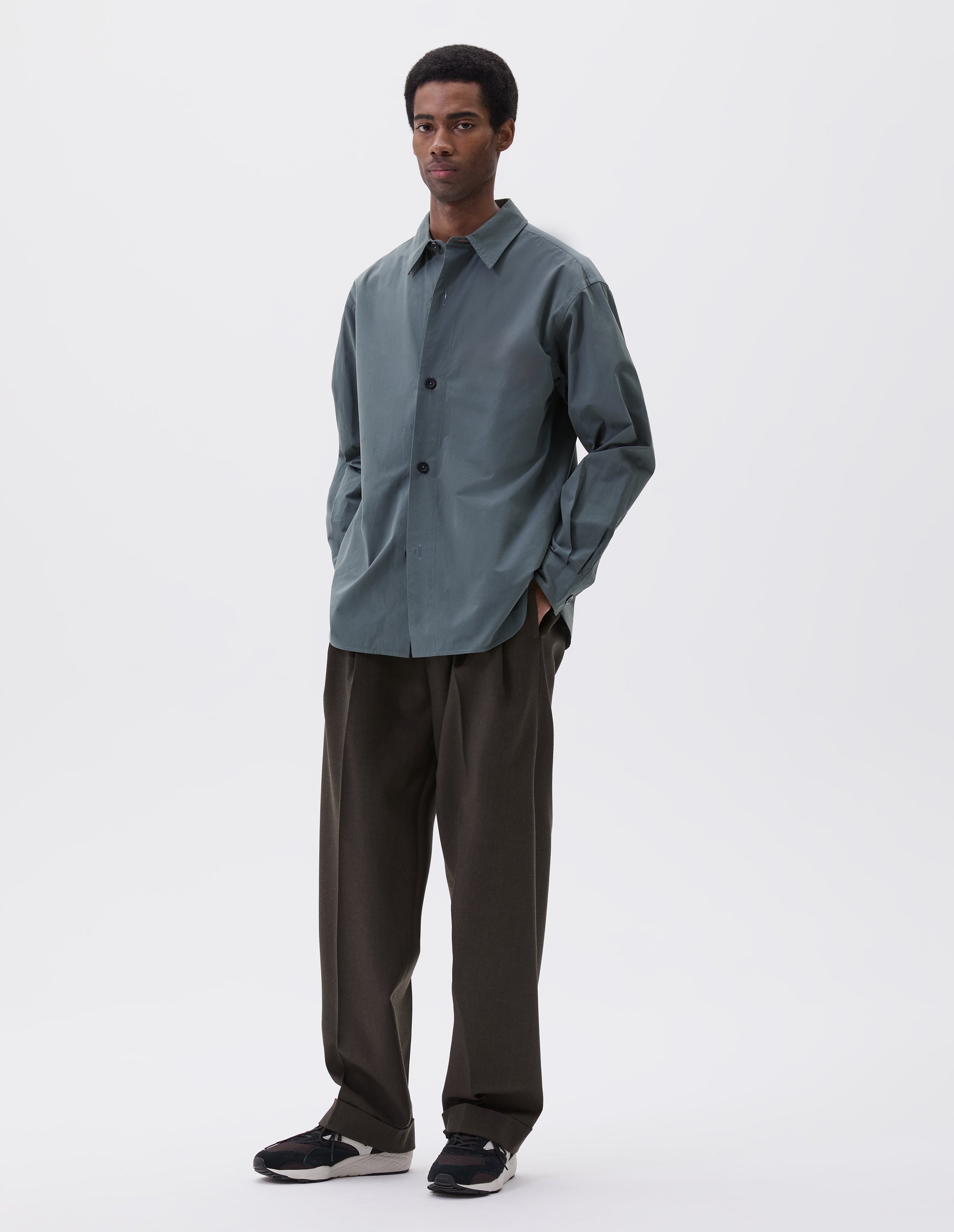 Full body image, front view of model wearing relaxed blue shirt with stitch through placket and horn buttons. Styled with khaki green trousers and black, brown and white trainers.