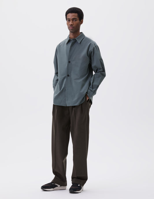 Full body image, front view of model wearing relaxed blue shirt with stitch through placket and horn buttons. Styled with khaki green trousers and black, brown and white trainers.