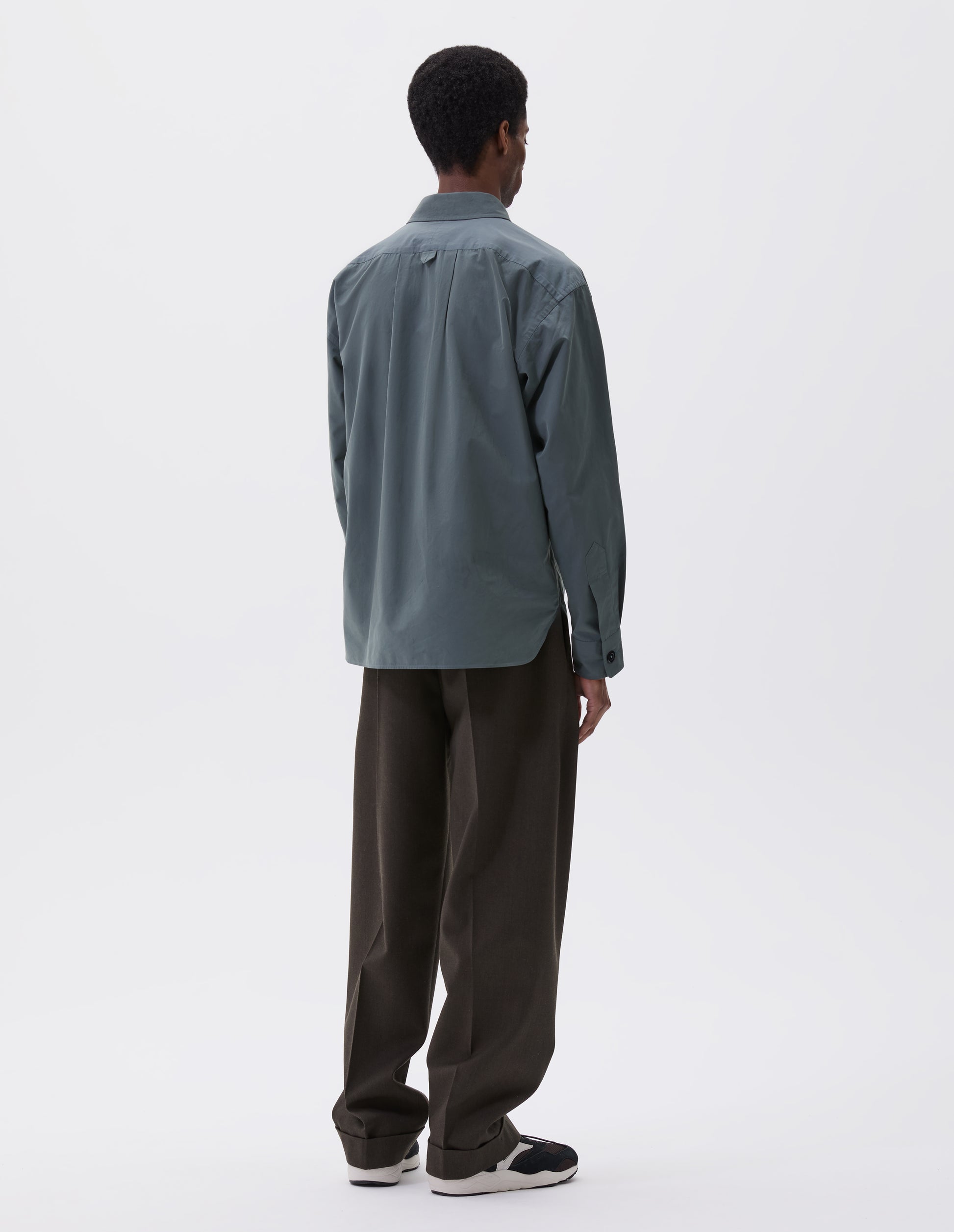 Full body image, back view of model wearing relaxed blue shirt with hanger loop detail on back, split yoke and horn buttons on cuffs. Styled with khaki green trousers and black, brown and white trainers.