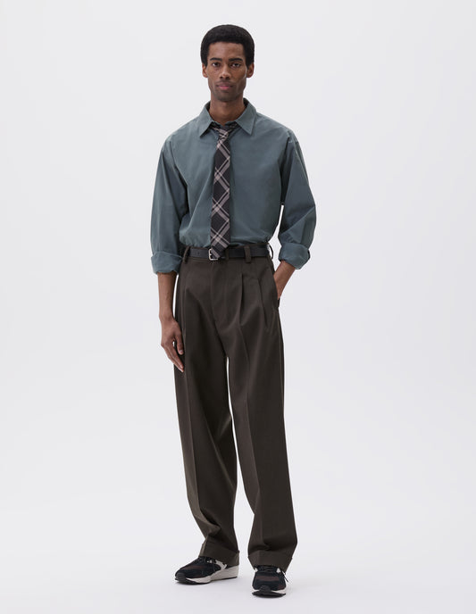 Full body image, front view of model wearing brownish green wool trousers with deep double pleat, horn button waistband and zip closure, belt loops and concealed side pockets. Styled blue shirt, green checked tie, black leather belt and black and brown trainers. 
