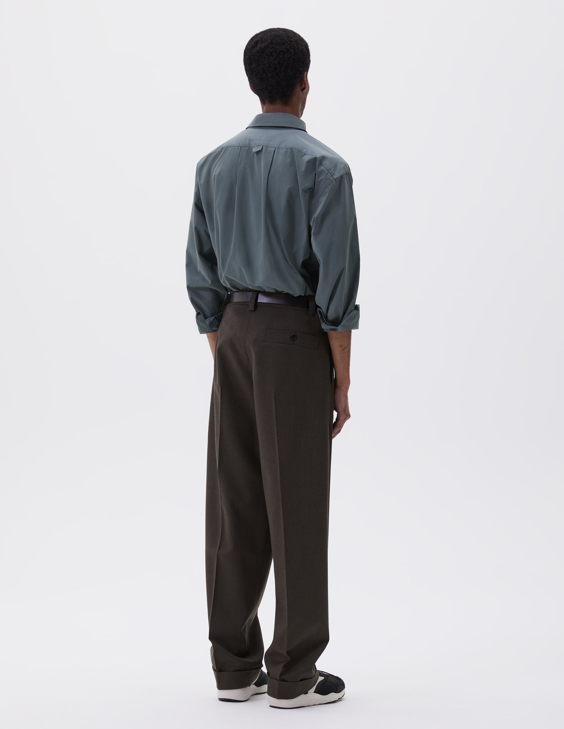 Full body image, back view of model wearing brownish green wool trousers with concealed side pockets and deep button through welt back pocket. Styled blue shirt, black leather belt and black and brown trainers.