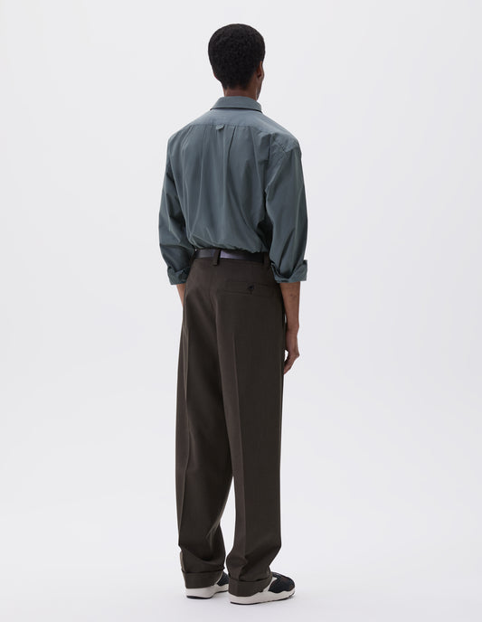 Full body image, back view of model wearing brownish green wool trousers with concealed side pockets and deep button through welt back pocket. Styled blue shirt, black leather belt and black and brown trainers.