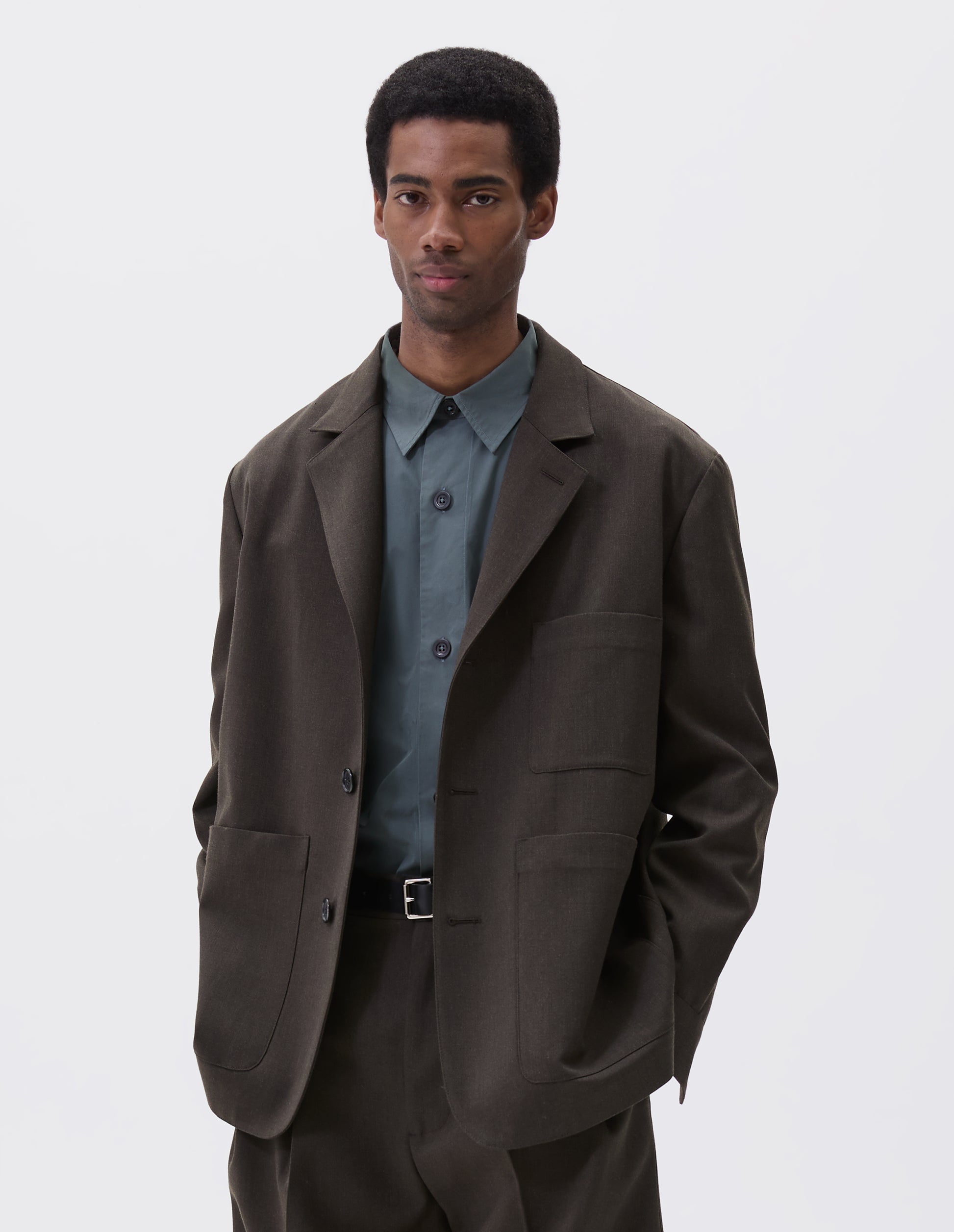 Cropped body image, front view of model wearing brownish green wool blazer with horn button fastening, deep wrap closure, shirt cuff sleeves and patch pockets. Styled over blue shirt and matching brownish green trousers. With black leather belt.