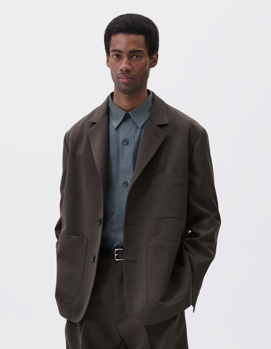 Cropped body image, front view of model wearing brownish green wool blazer with horn button fastening, deep wrap closure, shirt cuff sleeves and patch pockets. Styled over blue shirt and matching brownish green trousers. With black leather belt.
