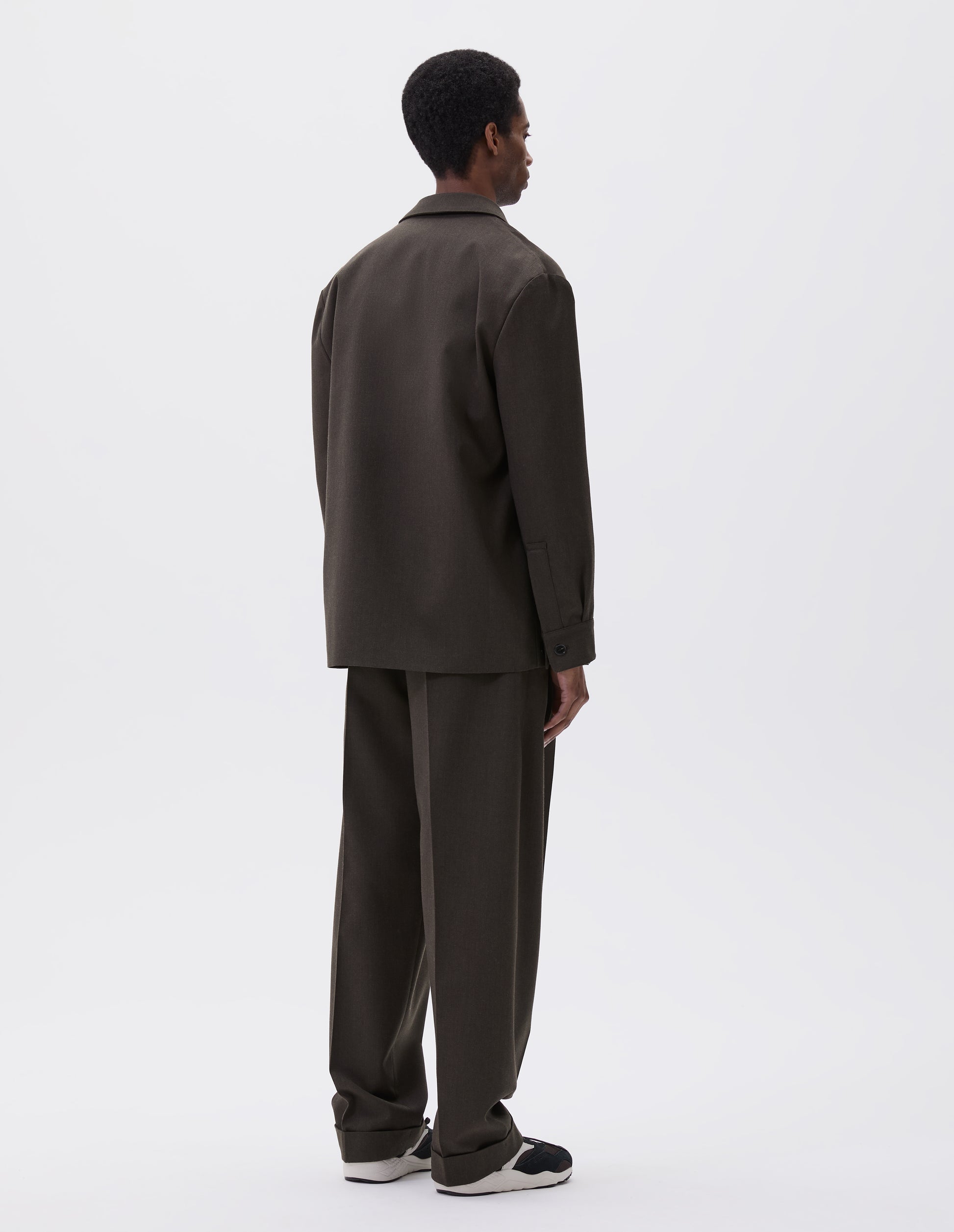 Full body image, back view of model wearing brownish green wool blazer with horn button shirt cuffs. Styled with matching brownish green trousers and black and brown trainers.