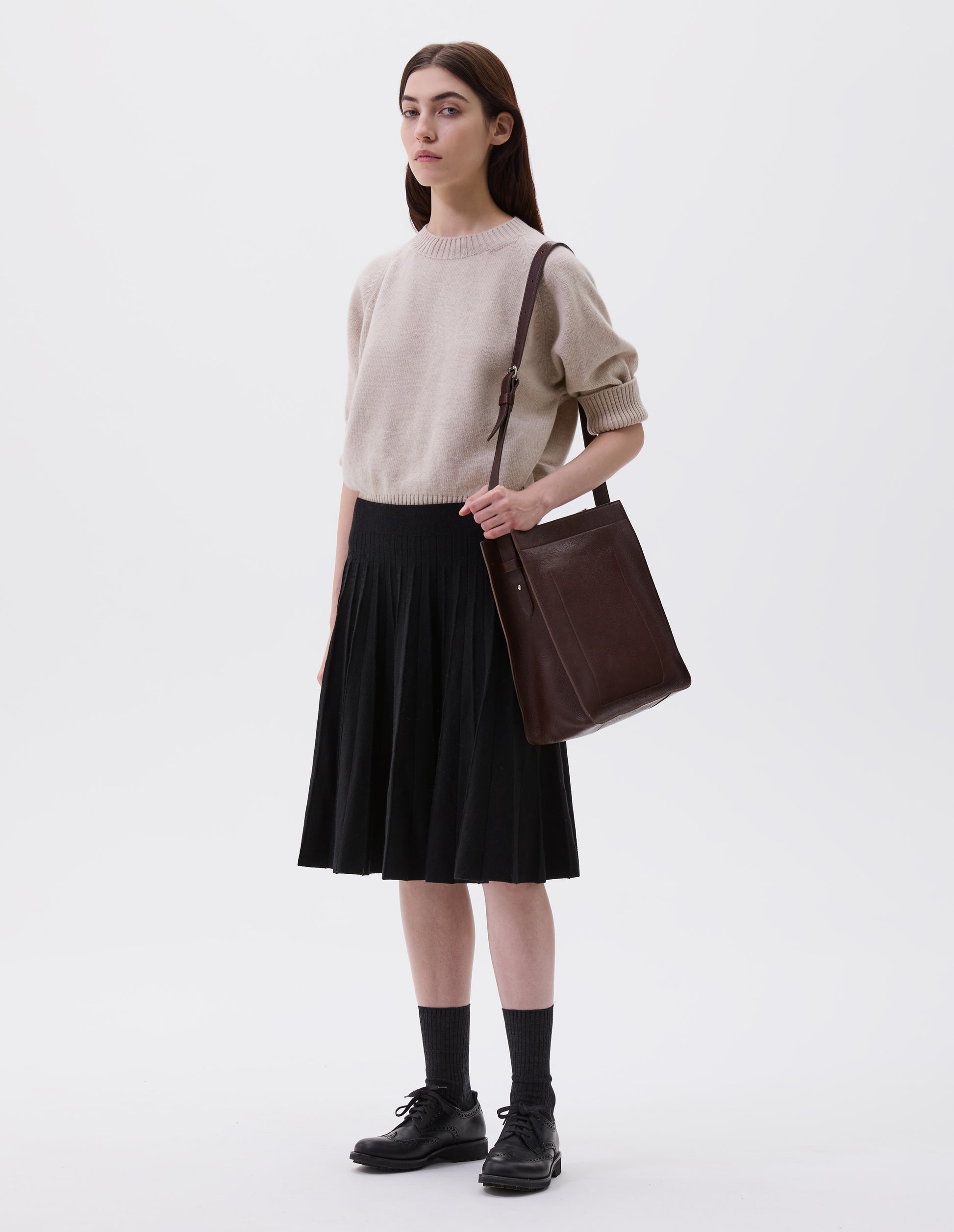 Full body image, side view of model wearing brown fine grain leather tote bag with a roller buckle adjustable shoulder strap and hand-stained turned-out seams. Styled with beige jumper, black knitted skirt, black socks and black leather brogues.