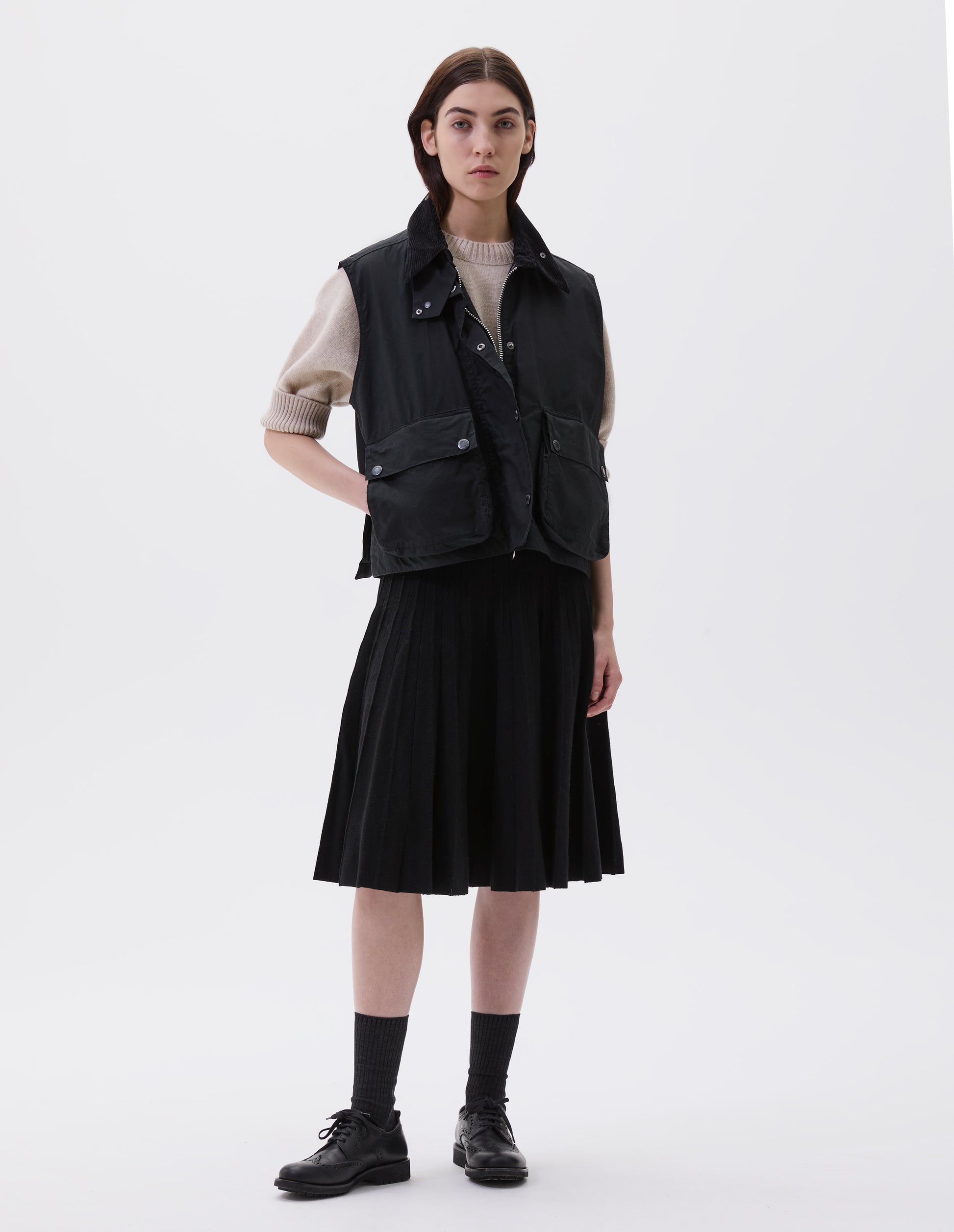 Full body image, front view of model wearing black boxy gilet with storm tab on corduroy collar, two-way zip and press stud fastening storm flap, large bellows pockets with press stud fastening envelope closure and concealed side pockets. Styled over beige jumper  with black pleated skirt, black socks and black leather brogues.