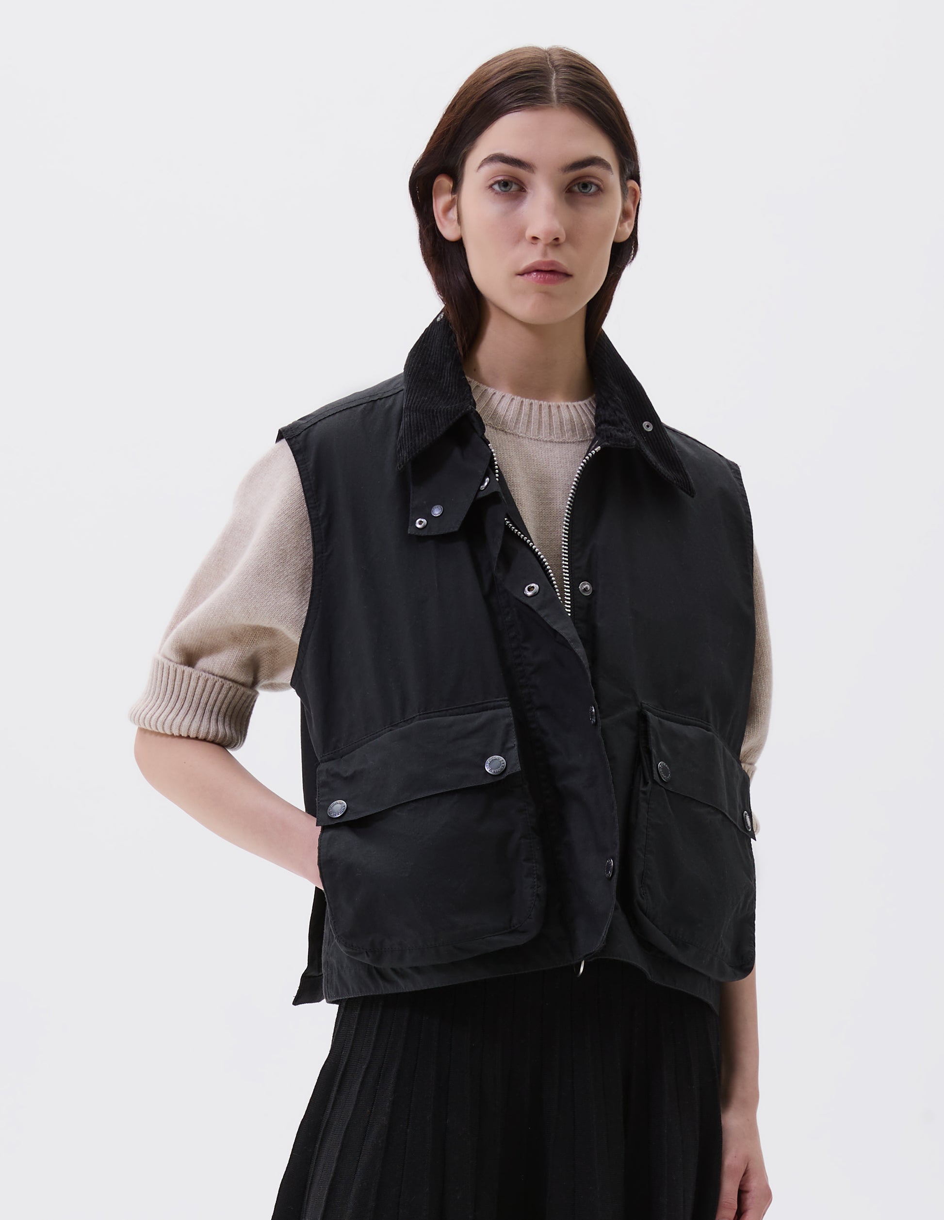 Cropped image, front view of model wearing black boxy gilet with storm tab on corduroy collar, two-way zip and press stud fastening storm flap, large bellows pockets with press stud fastening envelope closure and concealed side pockets. Styled over beige jumper  with black pleated skirt.