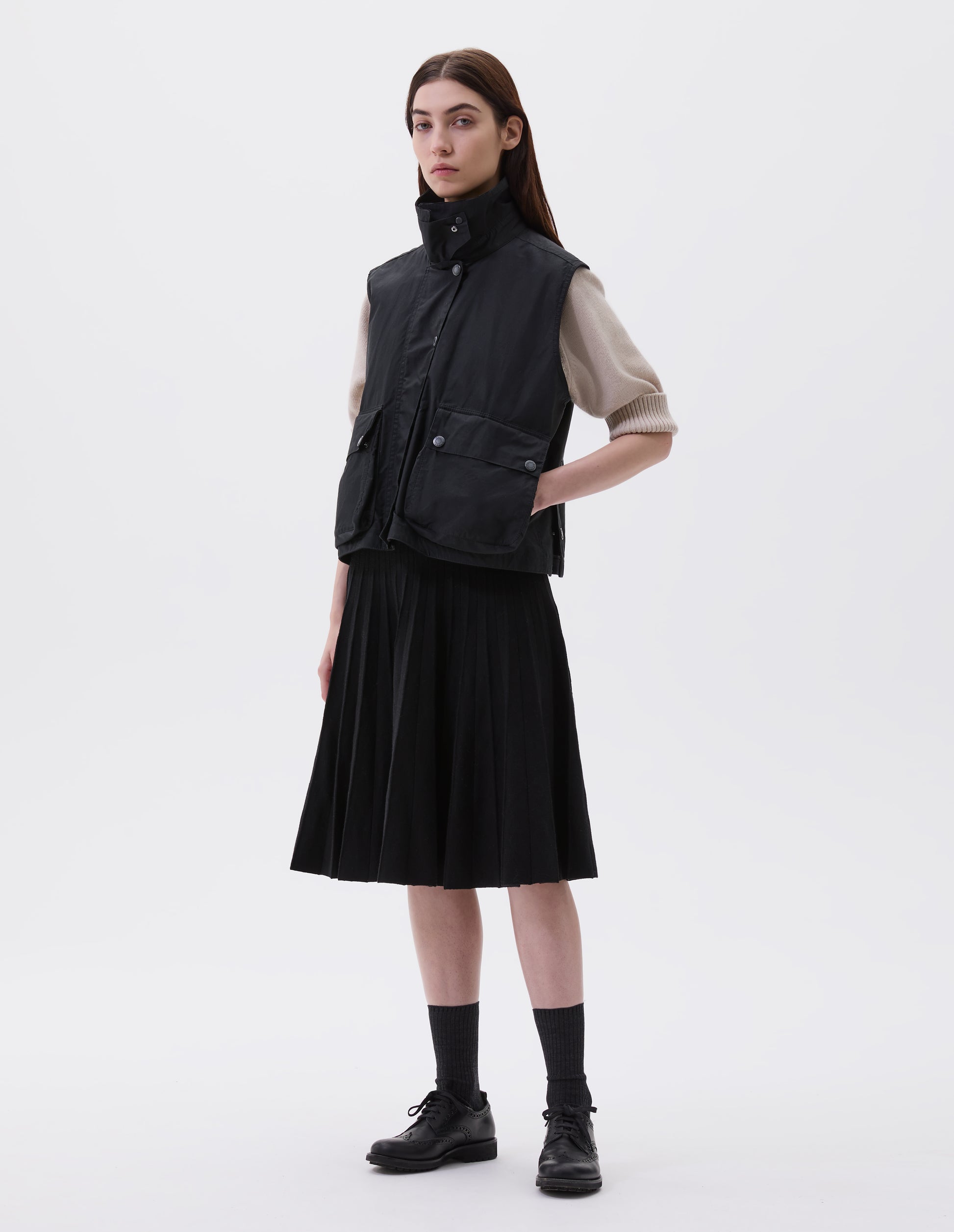 Full body image, front view of model wearing black boxy gilet with storm tab on stand collar, press stud fastening storm flap, large bellows pockets with press stud fastening envelope closure and concealed side pockets. Styled over beige jumper with black pleated skirt, black socks and black leather brogues.