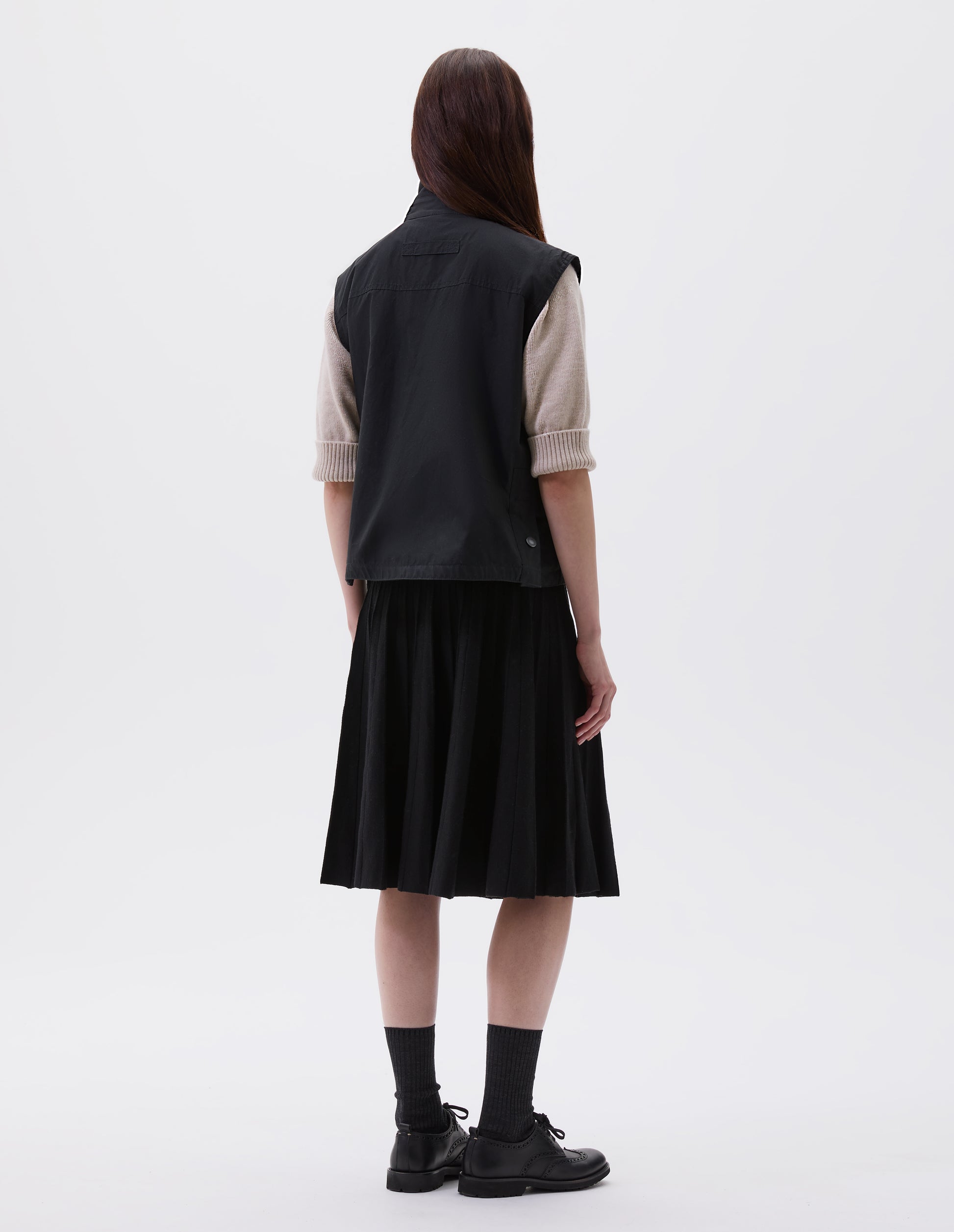 Full body image, back view of model wearing black boxy gilet with stand collar, hanger loop on back and press stud fastening vent split hem with gussets. Styled over beige jumper  with black pleated skirt, black socks and black leather brogues.