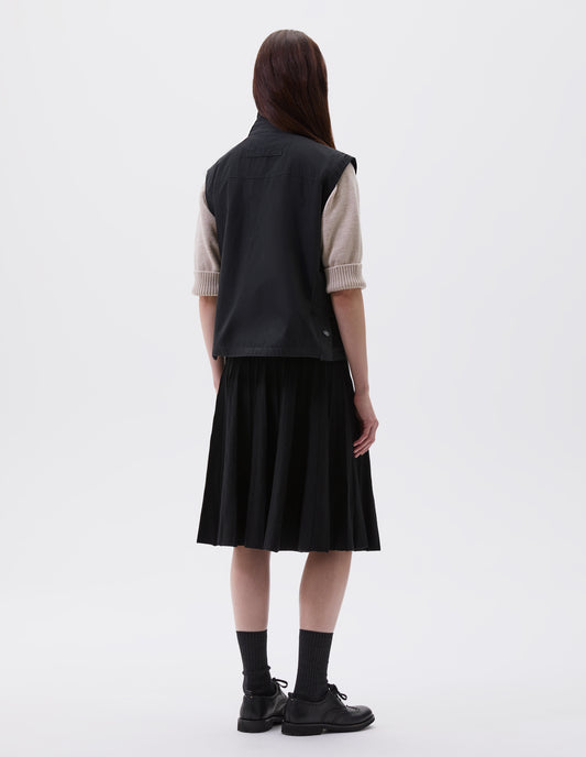Full body image, back view of model wearing black boxy gilet with stand collar, hanger loop on back and press stud fastening vent split hem with gussets. Styled over beige jumper  with black pleated skirt, black socks and black leather brogues.