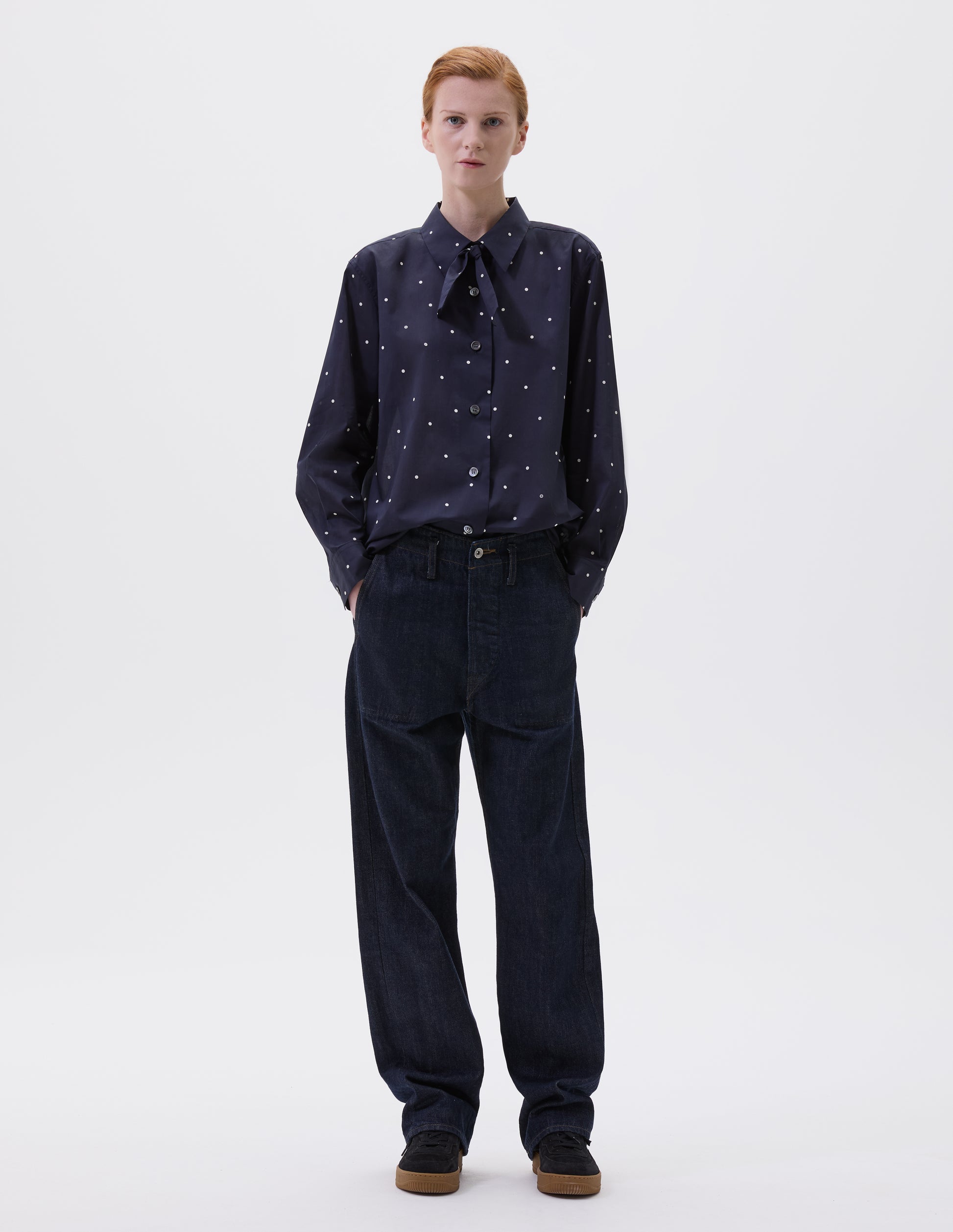 Full body image, front view of model wearing navy and white polka dot long sleeve shirt with wide stitch through placket, mother of pearl buttons, elongated back and removeable self fabric tie. Styled with indigo blue jeans and black suede trainers.