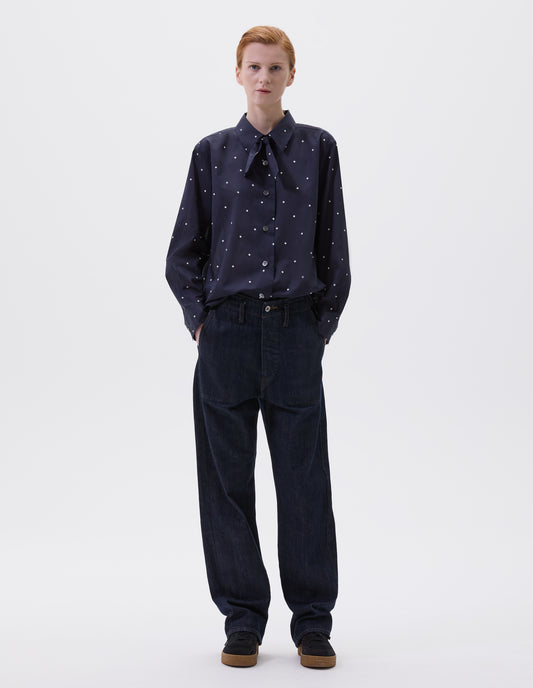 Full body image, front view of model wearing navy and white polka dot long sleeve shirt with wide stitch through placket, mother of pearl buttons, elongated back and removeable self fabric tie. Styled with indigo blue jeans and black suede trainers.
