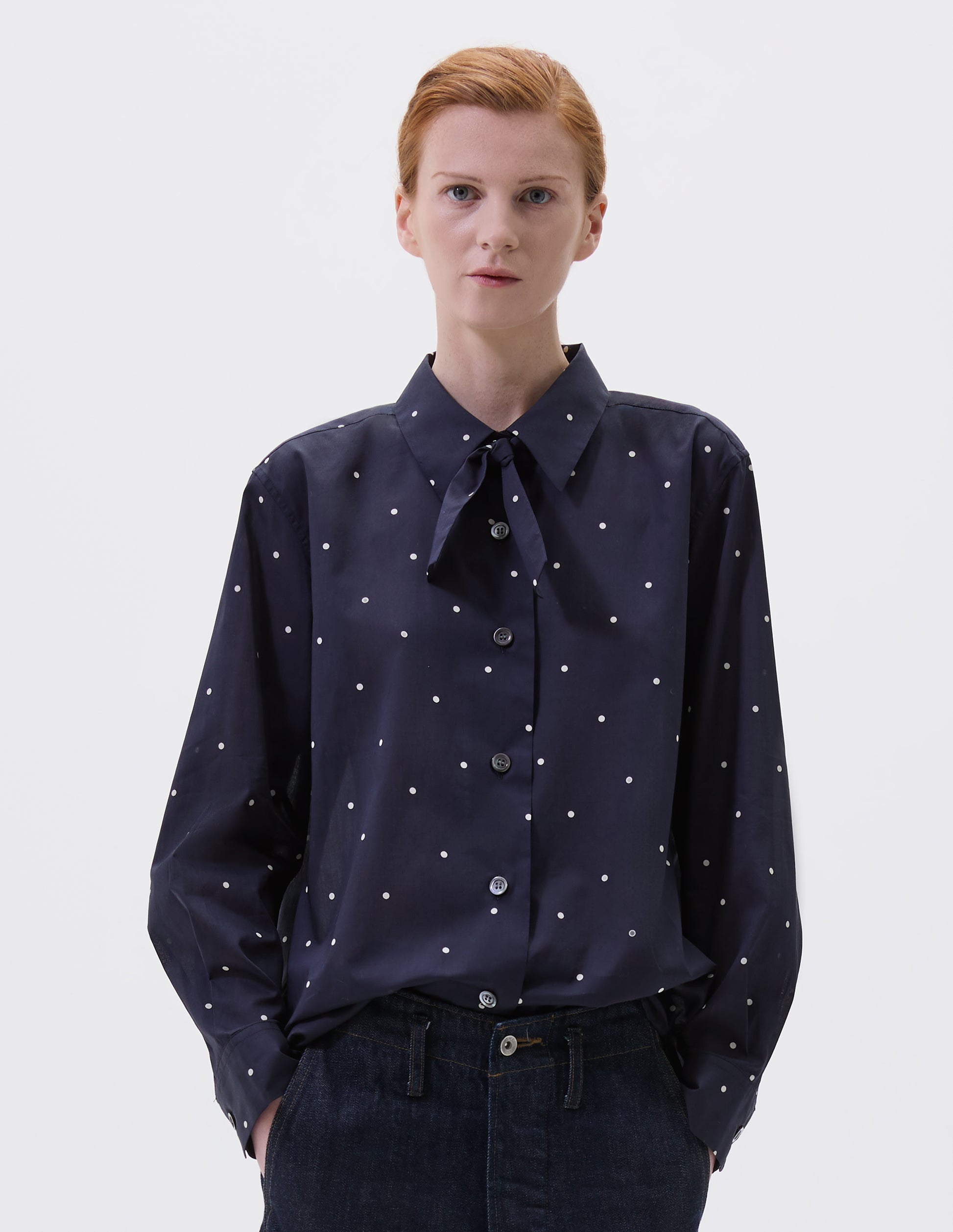 Cropped body image, front view of model wearing navy and white polka dot long sleeve shirt with wide stitch through placket, mother of pearl buttons, elongated back and removeable self fabric tie. Styled with indigo blue jeans.