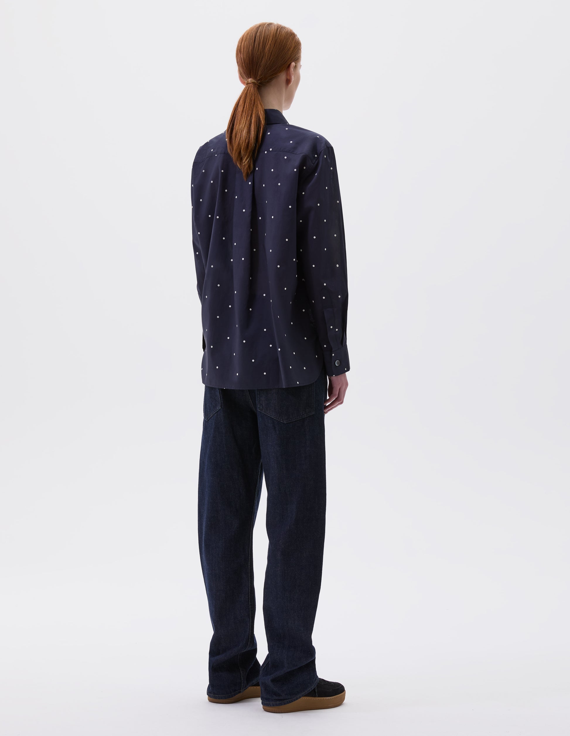 Full body image, back view of model wearing navy and white polka dot long sleeve shirt with elongated back and removeable self fabric tie. Styled with indigo blue jeans and black suede trainers.