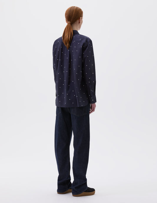Full body image, back view of model wearing navy and white polka dot long sleeve shirt with elongated back and removeable self fabric tie. Styled with indigo blue jeans and black suede trainers.