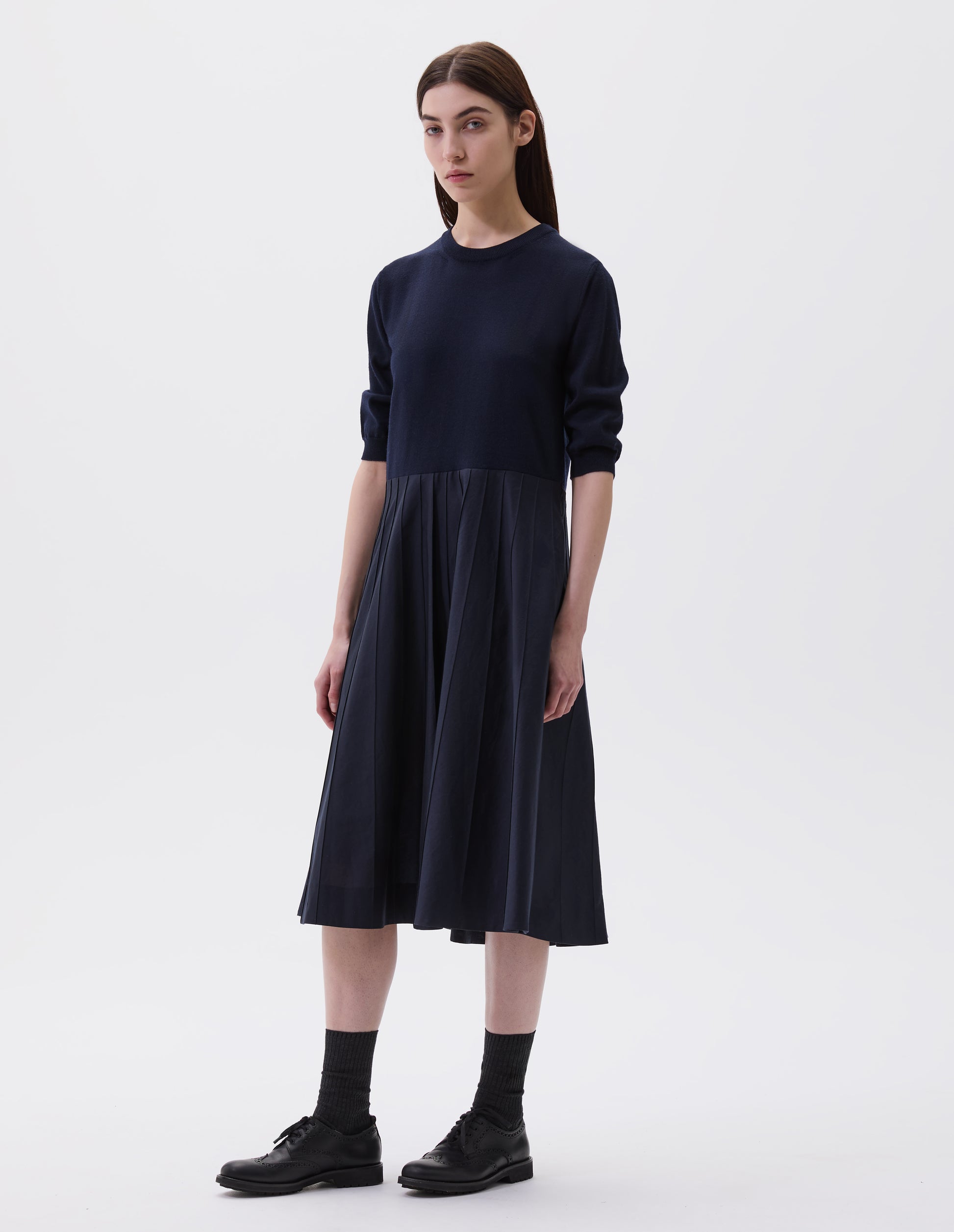 Full body image front view of model wearing navy blue short sleeve knitted and silk dress. Knitted merino wool top and cotton silk skirt. With concealed side pockets and pintuck detailing on skirt. Styled with black socks and black leather brogues.