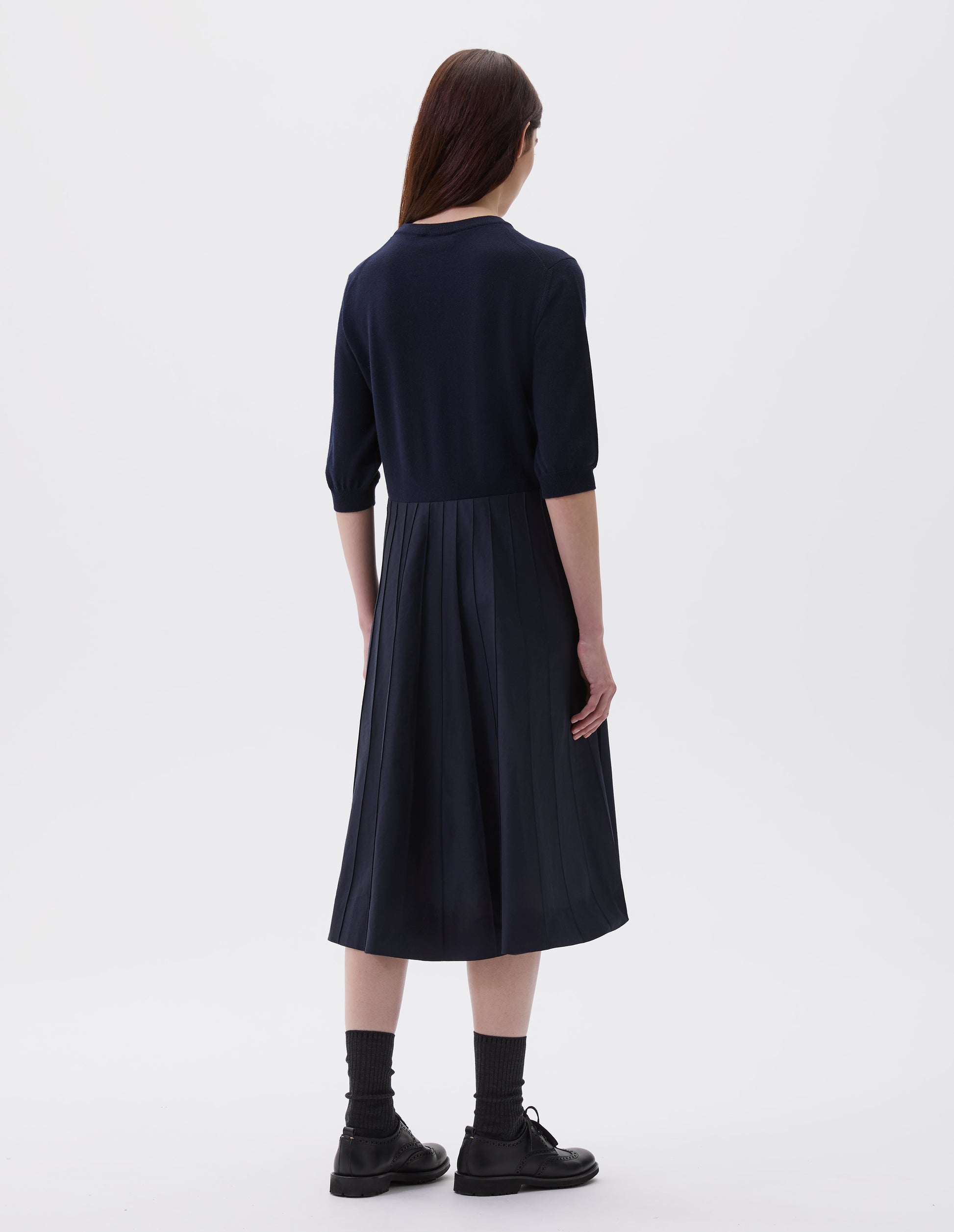 Full body image back view of model wearing navy blue short sleeve knitted and silk dress. Knitted merino wool top and cotton silk skirt. With concealed side pockets and pintuck detailing on skirt. Styled with black socks and black leather brogues.