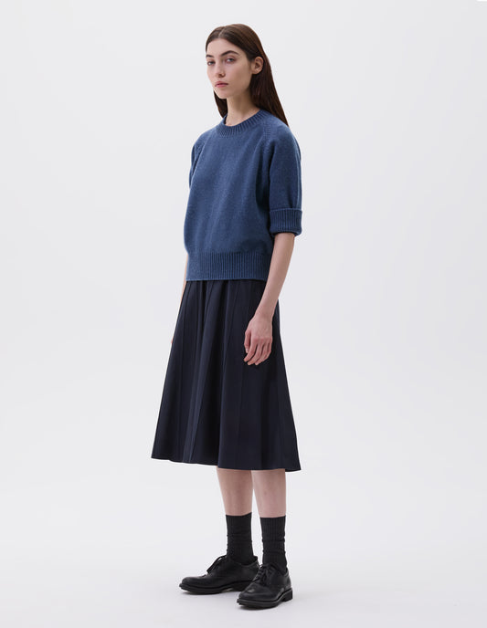 Full body image, front view of model wearing blue short sleeve crew neck jumper with raglan sleeves, wide ribbed hem and turned up wide ribbed cuffs, styled over dark blue knitted dress, black socks and black brogues.