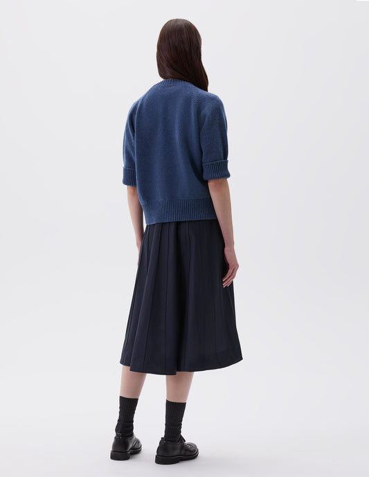 Full body image, back view of model wearing blue short sleeve crew neck jumper with raglan sleeves, wide ribbed hem and turned up wide ribbed cuffs, styled over dark blue knitted dress, black socks and black brogues.