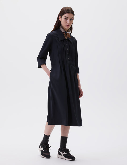 Full body image, front view of model with hand in pocket, wearing dark navy collared dress with pleat detailing, horn button closure and three quarter length sleeves. Styled with multicoloured scarf, black socks and black and brown trainers. 