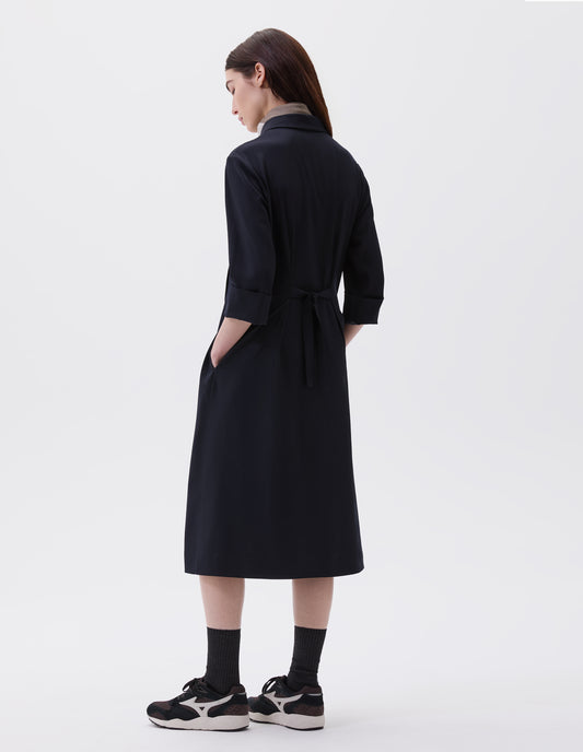 Full body image, back view of model with hands in pockets, wearing dark navy collared dress with tie closure on back and three quarter length sleeves. Styled with multicoloured scarf, black socks and black and brown trainers. 
