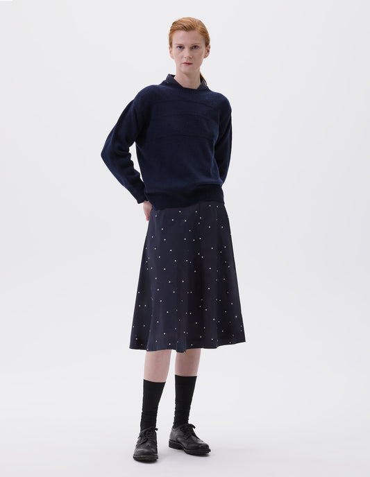 Full body image, front view of model wearing navy blue and white polka dot flared skirt, with concealed side pockets. Styled with matching shirt, navy blue jumper, long black socks and black leather brogues.
