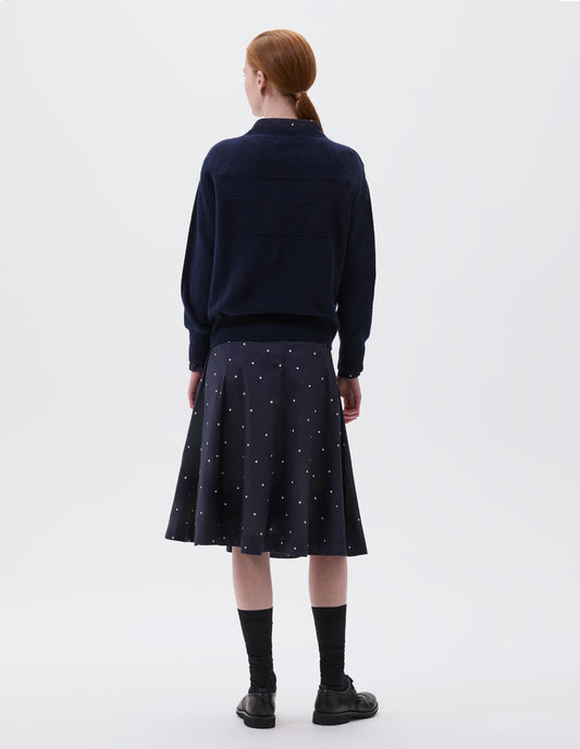 Full body image, back view of model wearing navy blue crew neck jumper with textured panelling on shoulders and back and contrasting rib on neck, cuffs and hem. Styled with navy and white polka dot shirt and skirt, black socks and black leather brogues.