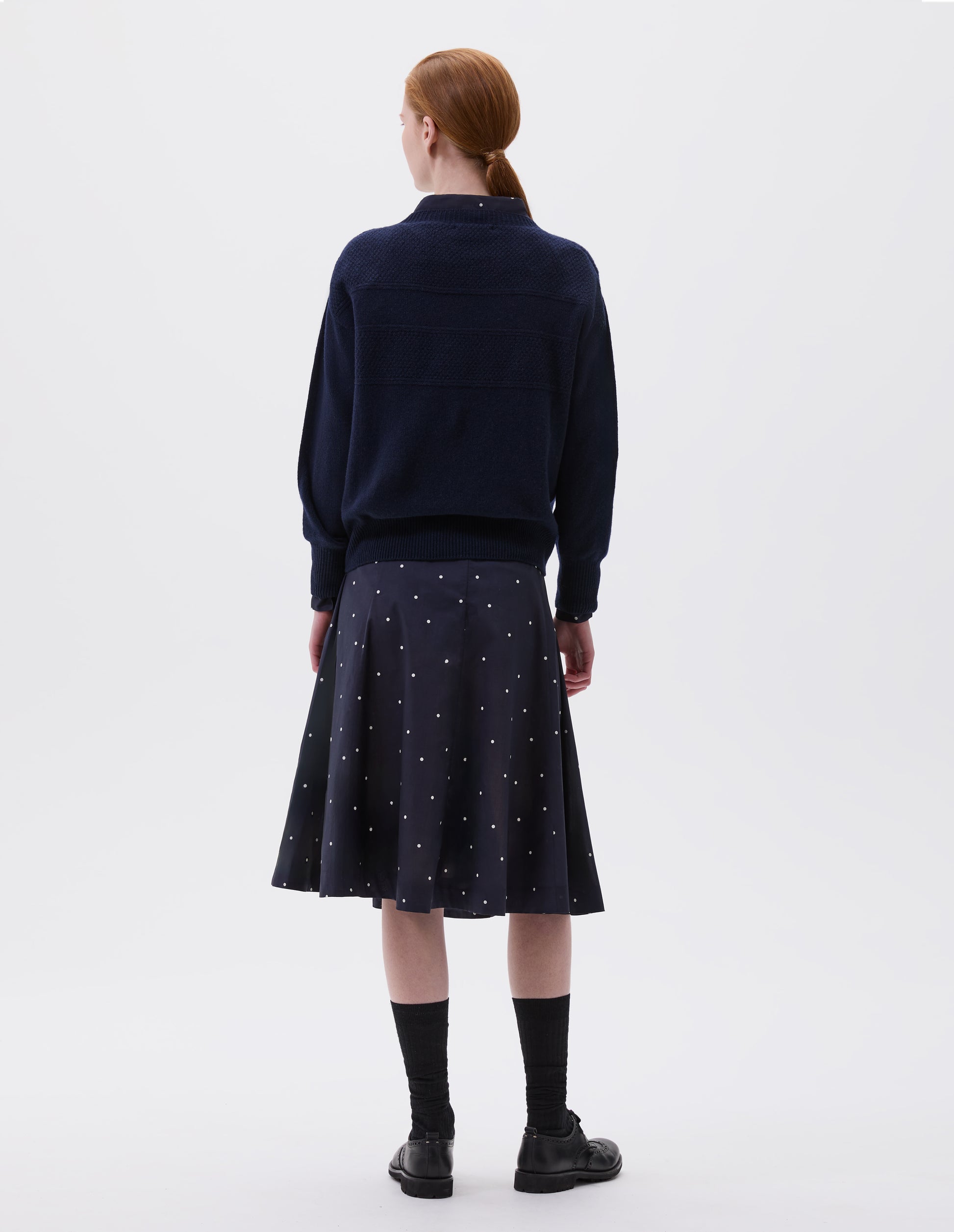 Full body image, back view of model wearing navy blue and white polka dot flared skirt, with concealed centre back zip fastening. Styled with matching shirt, navy blue jumper, long black socks and black leather brogues.
