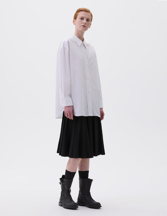 Full body image, front view of model wearing relaxed white shirt with wide raised placket and mother of pearl buttons, and turned up cuffs. Styled with black skirt, black socks and black leather biker boots.