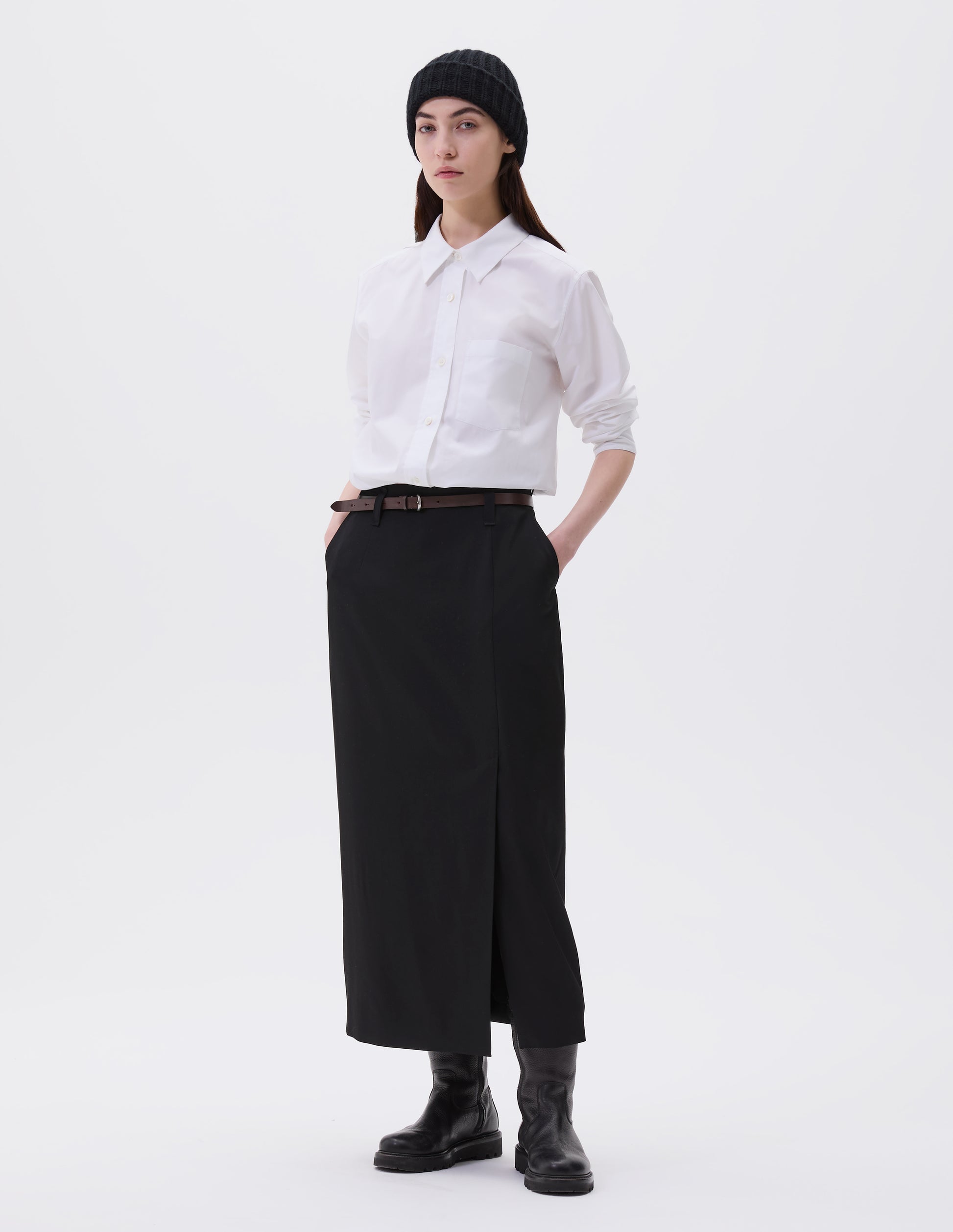 Full body image, front view of model wearing white shirt with raised placket and mother of pearl buttons, patch pocket on chest. Styled with black beanie, dark brown leather belt, black midi skirt and black leather boots.