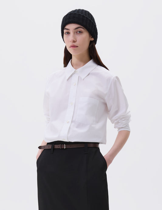 Cropped image, front view of model wearing white shirt with raised placket and mother of pearl buttons, patch pocket on chest. Styled with black beanie, dark brown leather belt and black skirt.