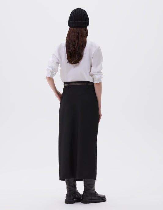 Full body image, back view of model wearing black fluid wool twill vent skirt, with belt loops. Styled with black wool beanie hat, white cotton shirt, brown slim leather belt and black leather boots.