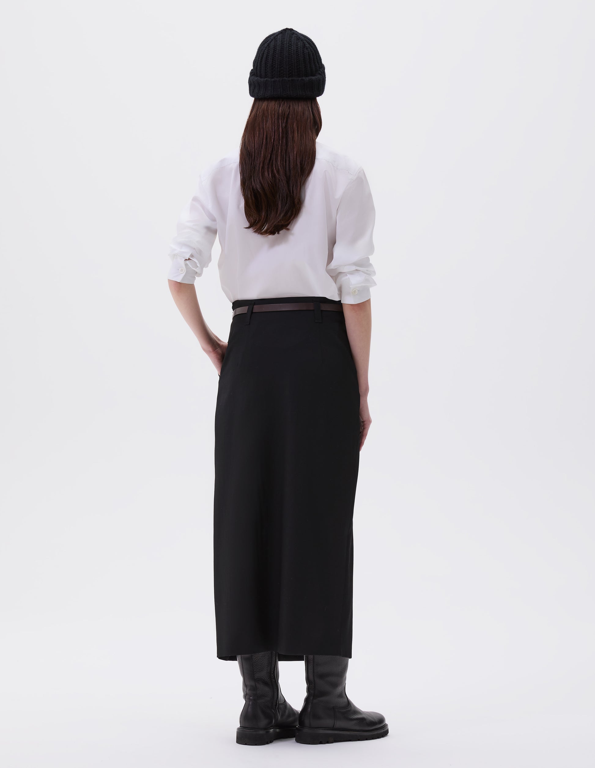 Full body image, back view of model wearing white shirt with mother of pearl buttons on cuffs. Styled with black beanie, dark brown leather belt, black midi skirt and black leather boots.