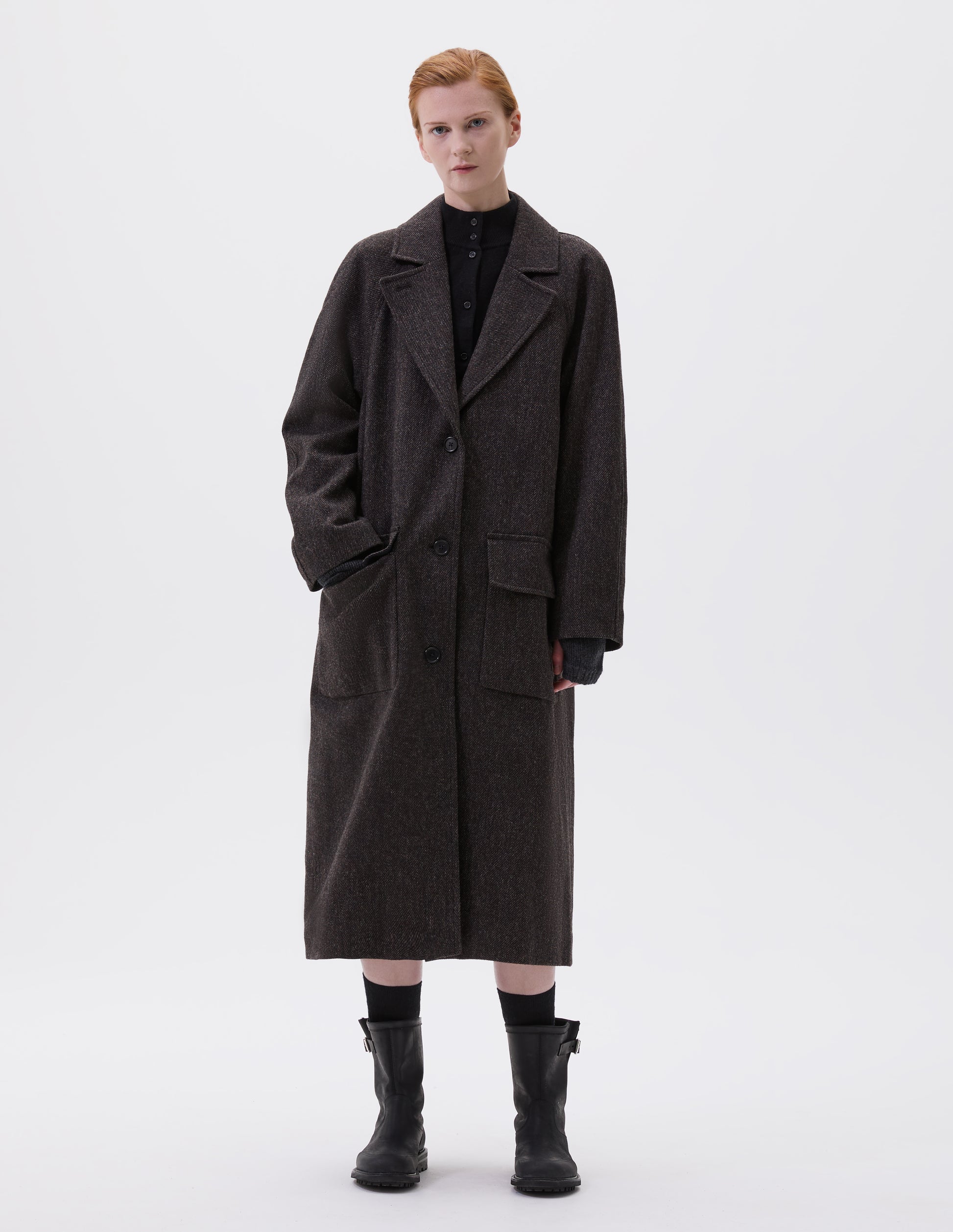 Full body image, front view of model wearing brown wool longline overcoat with raglan sleeve, button fastening lapel and deep flap patch pockets. Styled with black high neck cardigan, black socks and black leather boots. 