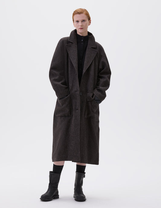 Full body image, front view of model wearing brown wool longline overcoat with raglan sleeve, button fastening lapel and deep flap patch pockets. Styled with black high neck cardigan, black socks and black leather boots. 