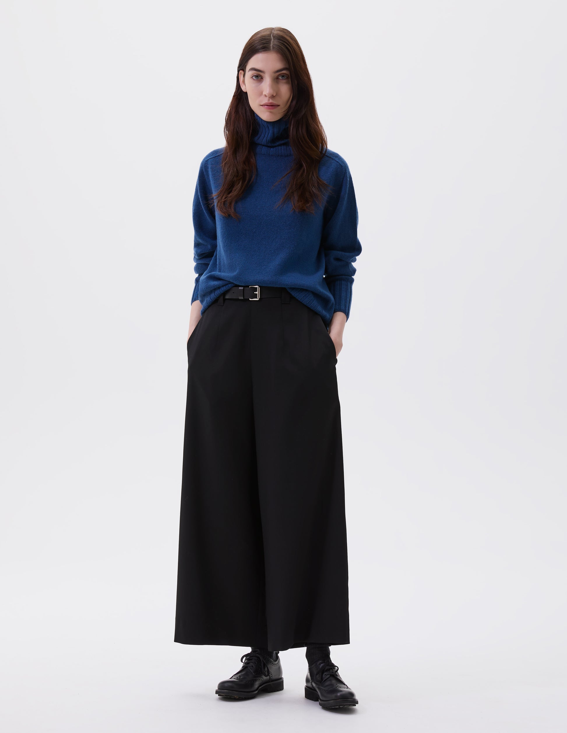 Full body image, front view of model wearing black wool wide leg culottes, with belt loops, side pockets and concealed zip fastening. Styled with blue turtleneck jumper, black leather belt and black leather brogues.