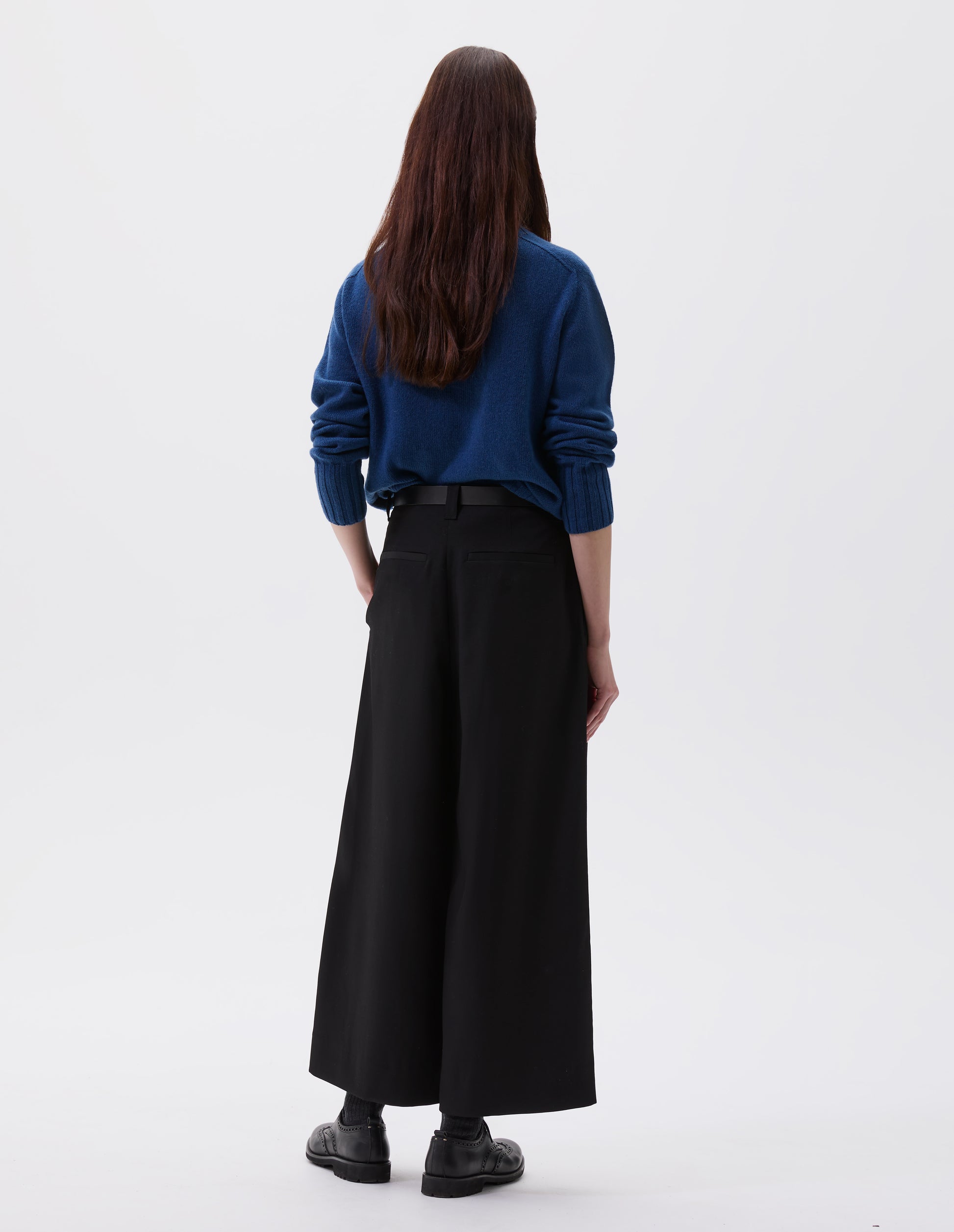 Full body image, back view of model wearing black wool wide leg culottes, with belt loops, side pockets and welt back pockets. Styled with blue turtleneck jumper, black leather belt and black leather brogues.