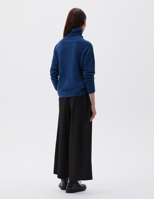 Full body image, back view of model wearing blue high neck cashmere jumper, with saddle sleeve and contrasting wide rib on neck, cuffs and hem. Styled with black culottes, black socks and black leather brogues.