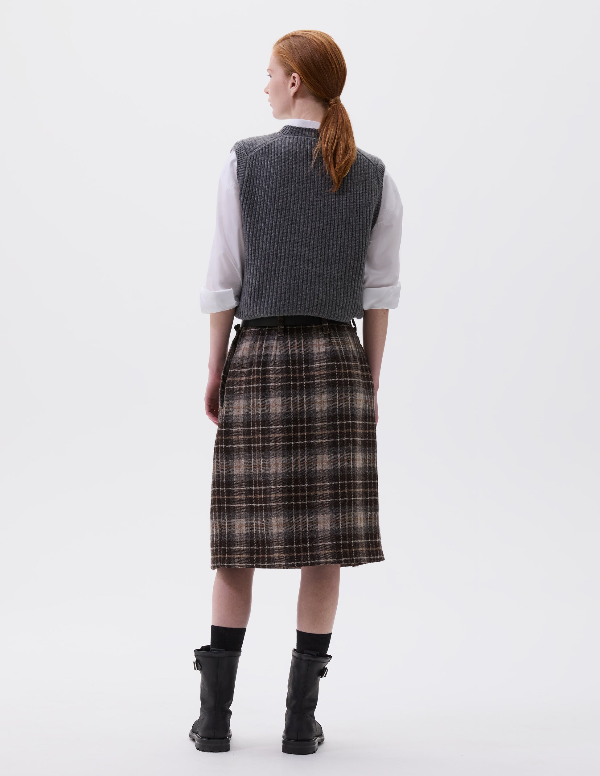 Full body image back view of model wearing beige and brown checked wool skirt. With deep pleated waistband, belt loops and concealed side pockets Styled with white shirt, grey sleeveless jumper, black leather belt, black socks and black leather boots.
