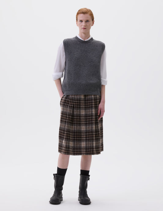 Full body image, front view of model wearing grey sleeveless merino wool slipover, with contrast rib on neck, armhole and hem. Styled with white shirt, brown and greyish checked skirt and black leather boots.