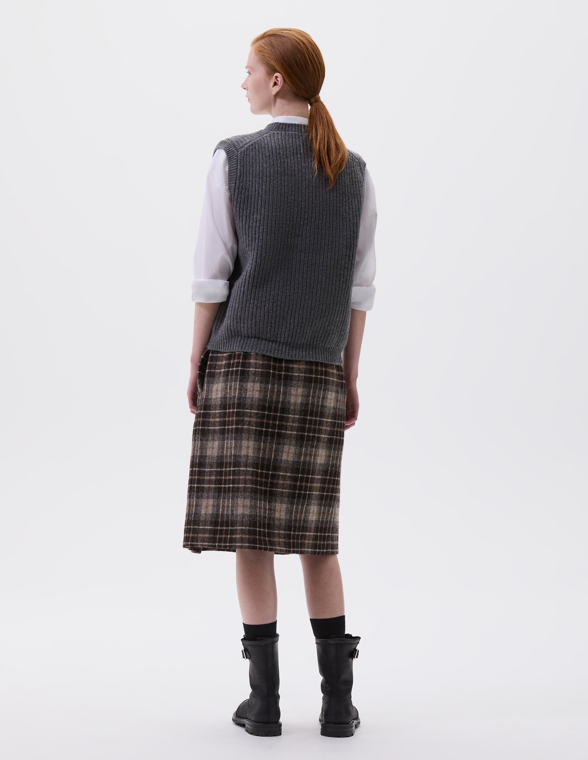 Full body image, back view of model wearing grey sleeveless merino wool slipover, with contrast rib on neck, armhole and hem. Styled with white shirt, brown and greyish checked skirt and black leather boots.