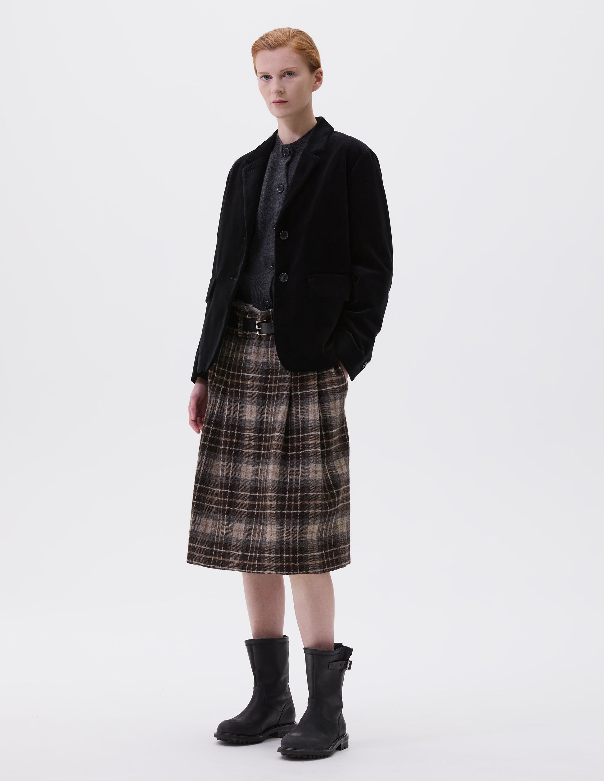 Full body image, front view of model wearing black blazer with horn buttons on lapel and cuffs. Styled over dark grey slipover and with brown tartan skirt, black leather belt and black leather biker boots.