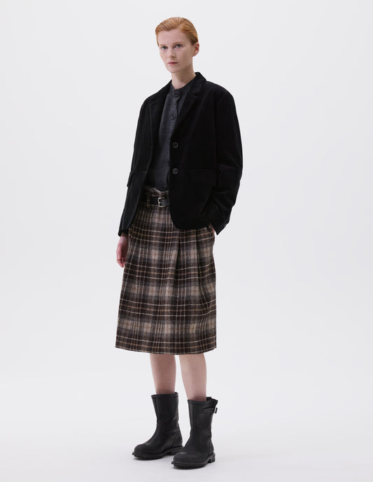 Full body image, front view of model wearing black blazer with horn buttons on lapel and cuffs. Styled over dark grey slipover and with brown tartan skirt, black leather belt and black leather biker boots.