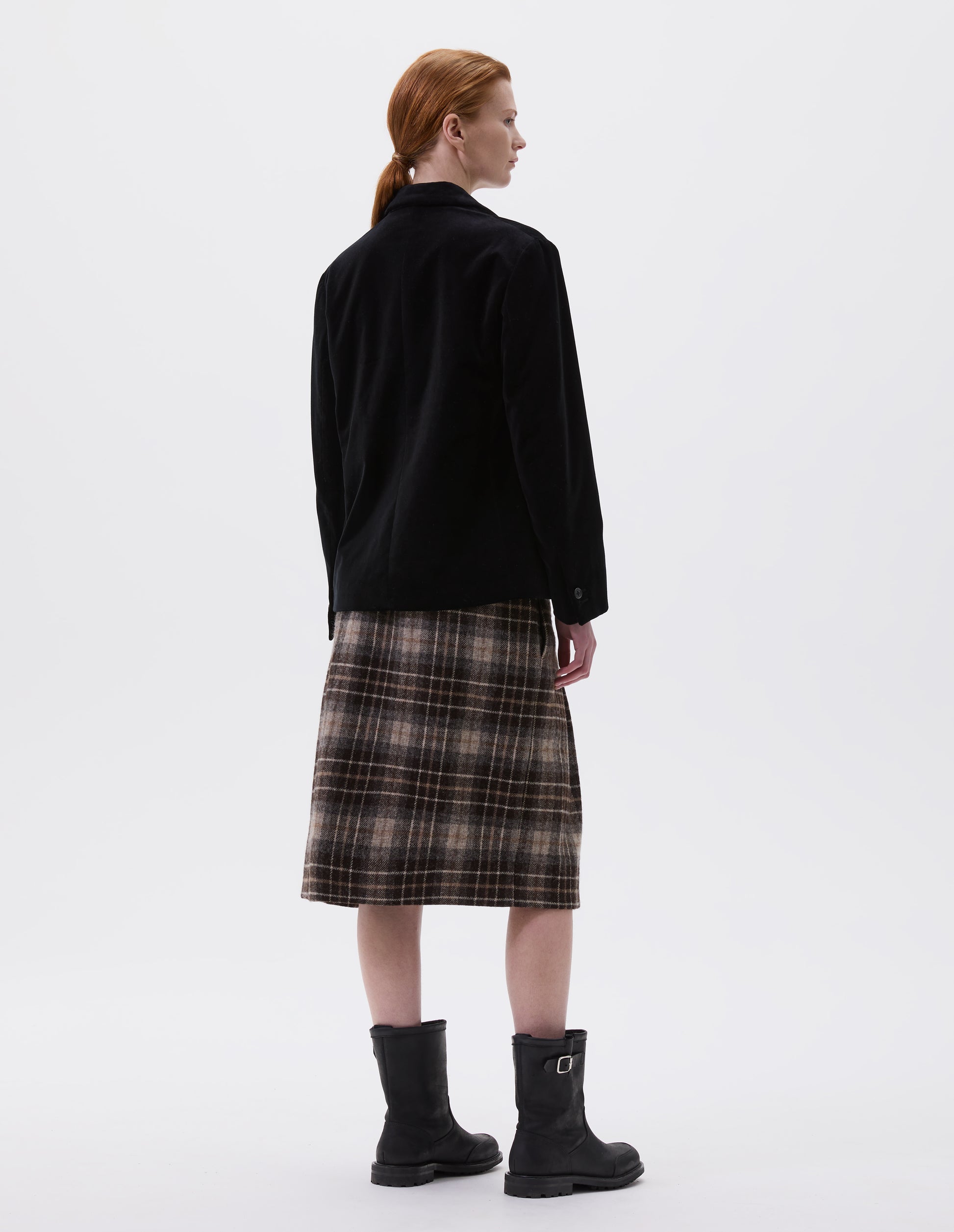Full body image, back view of model wearing black blazer with horn buttons on cuffs. Styled with brown tartan skirt and black leather biker boots.
