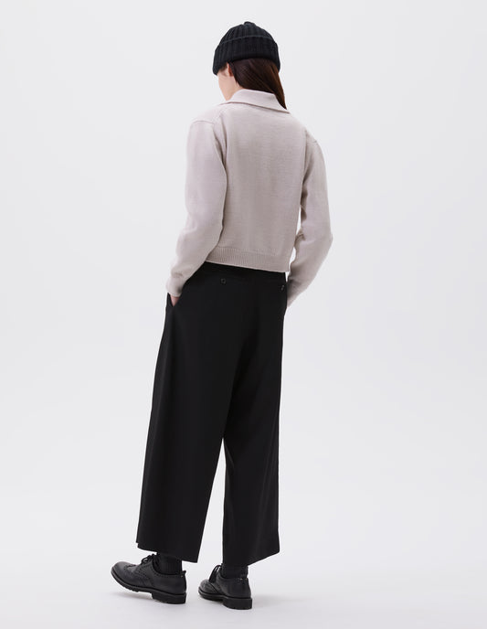 Full body image, back view of model wearing white boxy cropped cardigan with collar, black horn buttons and contrast rib on collar, cuffs and hem. Styled with black beanie, black culottes and black socks with black brogues.