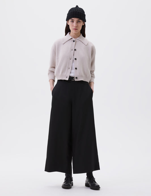 Full body image, front view of model wearing white boxy cropped cardigan with collar, black horn buttons and contrast rib on collar, cuffs and hem. Styled over white t-shirt with black beanie, black leather belt, black culottes, and black socks with black brogues.